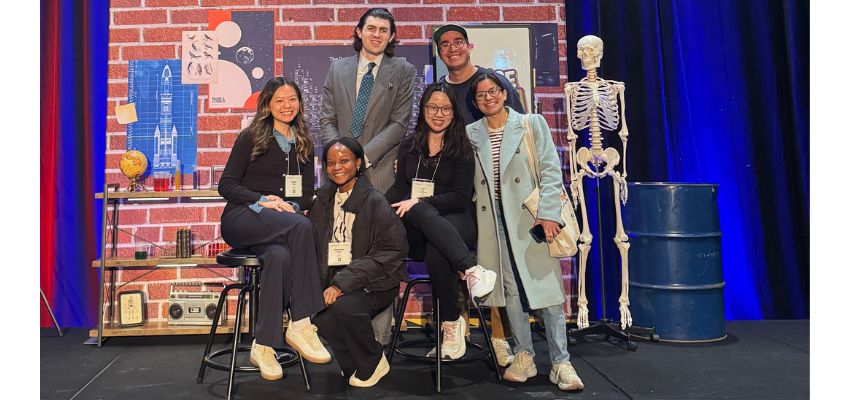 A group of students poses on a stage decorated with science-themed props, including a skeleton model, during a student event or competition.