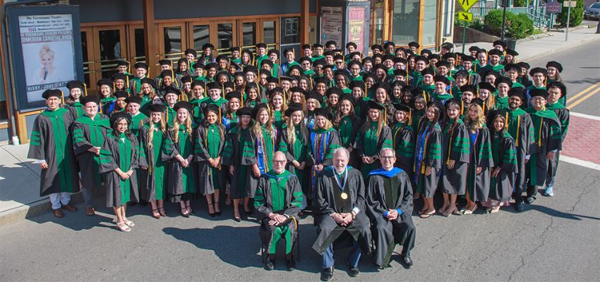 Members of the graduating class and faculty gather outside a theater for a formal class photo in academic regalia.