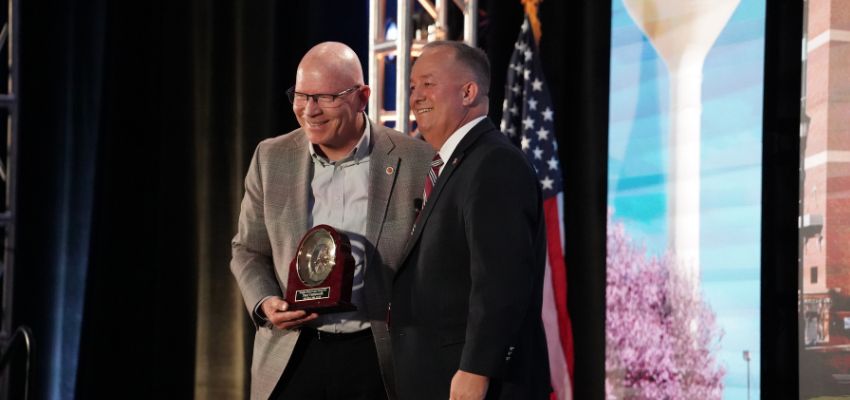 An award recipient accepts a commemorative plaque during a recognition ceremony while posing on stage with a presenter.