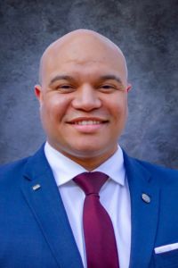 Professional headshot of a man in a blue suit and burgundy tie against a studio backdrop.