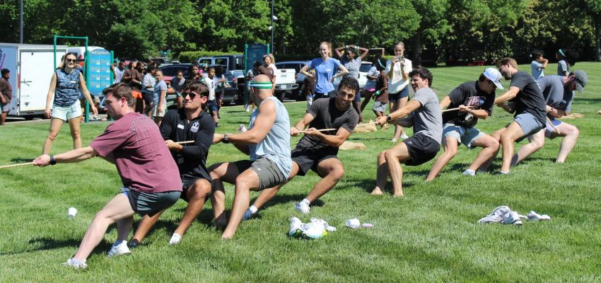 Medical students participate in a tug-of-war competition during an outdoor campus event while spectators watch nearby.