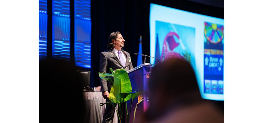 A speaker addresses attendees from a podium during a conference presentation, with a large screen displaying presentation materials in the background.