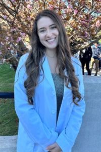 Portrait of a student wearing a white coat outdoors on campus, smiling in front of flowering trees.