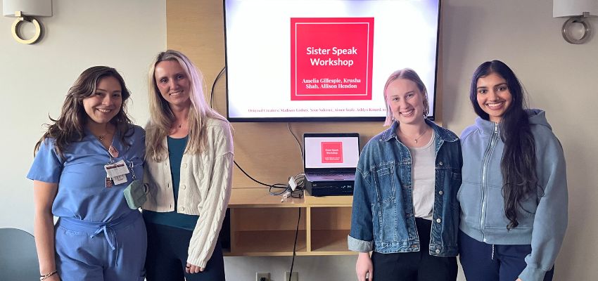 Four students stand together beside a presentation screen displaying “Sister Speak Workshop” during a student-led educational program.