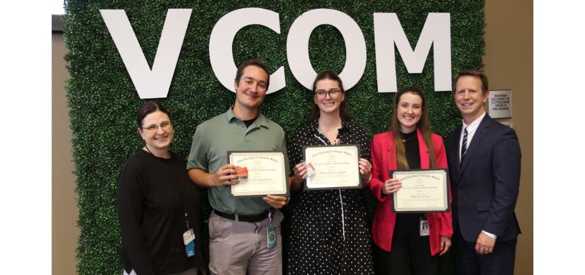 Three students holding certificates pose with two faculty or staff members in front of a VCOM-branded backdrop during an awards recognition ceremony.
