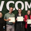 ThreestudentsholdingcertificatesposewithtwofacultyorstaffmembersinfrontofaVCOM-brandedbackdropduringanawardsrecognitionceremony