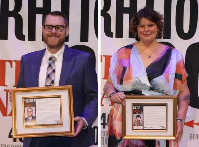 Two award recipients hold framed recognitions while posing on stage during an honors or alumni awards ceremony.