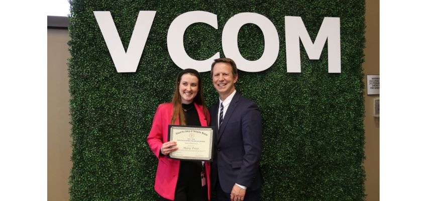 An award recipient poses with a faculty leader while holding a certificate in front of a VCOM-branded backdrop during a student recognition ceremony.