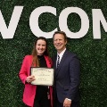 AnawardrecipientposeswithafacultyleaderwhileholdingacertificateinfrontofaVCOM-brandedbackdropduringastudentrecognitionceremony