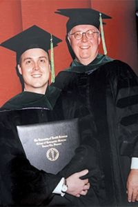 A graduate in academic regalia poses with a faculty member while holding a diploma during a commencement ceremony.