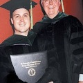 Agraduateinacademicregaliaposeswithafacultymemberwhileholdingadiplomaduringacommencementceremony