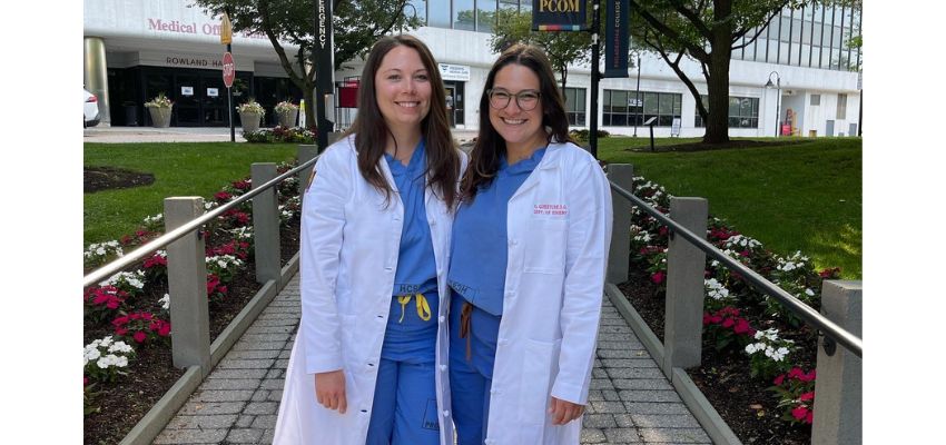 Two medical residents in white coats stand together outside a hospital entrance on the PCOM campus.