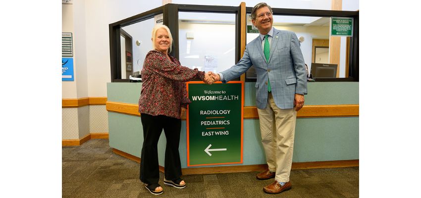 Two leaders shake hands beside a new directional sign inside a WVSOM Health facility during a ribbon-cutting or dedication event.