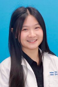Professional headshot of a medical student wearing a white coat against a blue studio background.