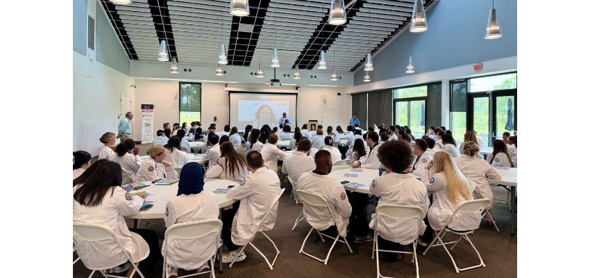 Medical students in white coats attend an orientation or educational session in a classroom while a speaker presents at the front of the room.