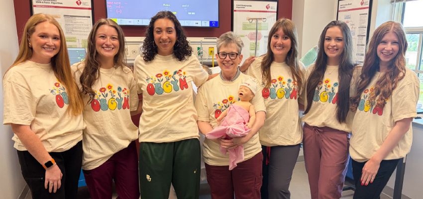 Medical students and a faculty member wearing matching OB/GYN shirts pose together in a clinical simulation lab with an infant manikin.