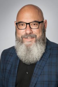 Portrait of a TCOM faculty or staff member with a long gray beard wearing glasses and a blue plaid blazer.