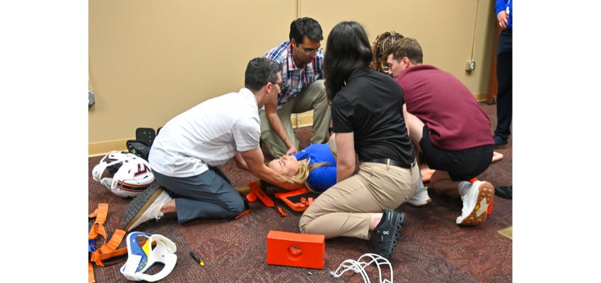 Medical students work together during an emergency response simulation, treating a patient lying on the floor with cervical stabilization equipment nearby.