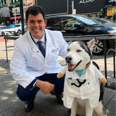A medical student kneels beside a Siberian Husky wearing a white doctor's coat during a community outreach event.