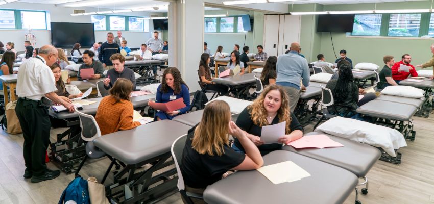 Medical students and faculty work in small groups around examination tables during a hands-on classroom session.