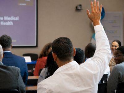 Audience member raises his hand during a presentation as other attendees sit facing the speaker.