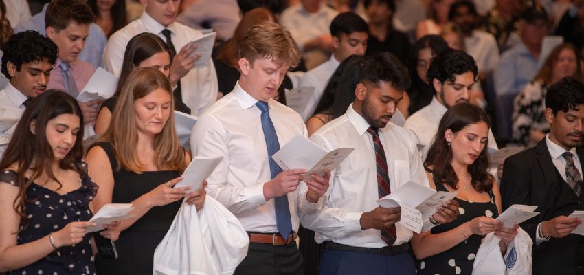 Students stand together reading from printed programs during a formal ceremony.