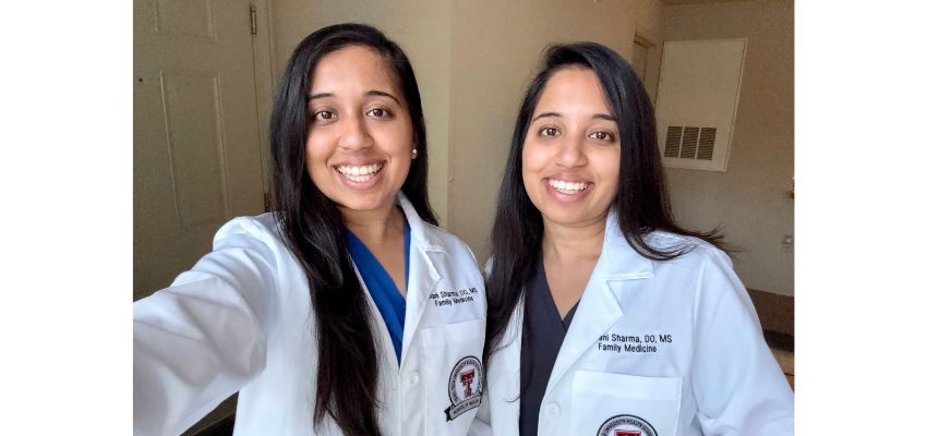Two physicians in white coats smile for a selfie while wearing medical scrubs.