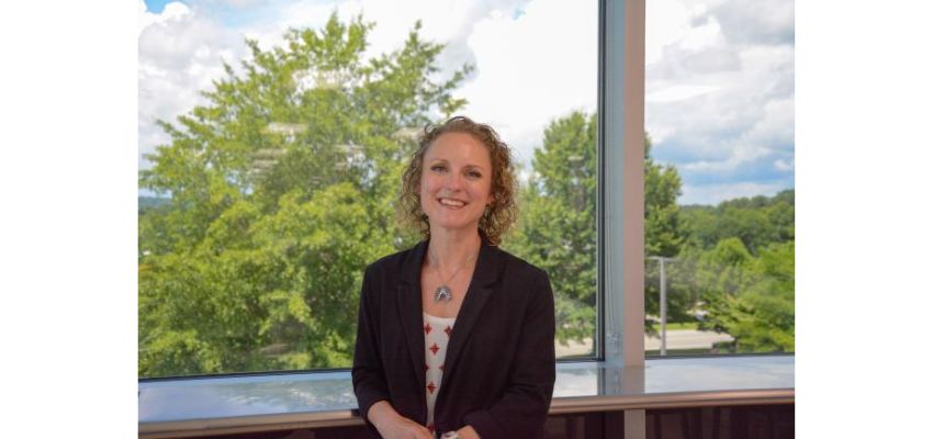 Portrait of a smiling faculty member standing indoors in front of large windows overlooking a wooded landscape.