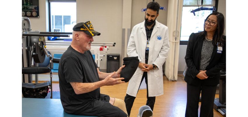 A veteran with a prosthetic leg speaks with two healthcare professionals in a rehabilitation facility.