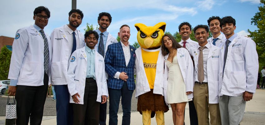 Medical students in white coats pose with a faculty member and the Rowan University owl mascot outdoors.