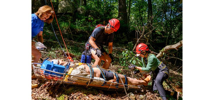 Rescue personnel wearing helmets and climbing gear transport a person secured to a rescue litter through a wooded area.