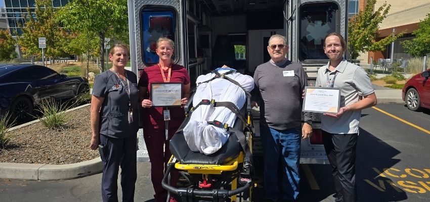 Four people pose beside an ambulance and a stretcher, with two holding certificates.