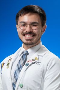 Portrait of a smiling medical student in a white coat and tie against a blue background.