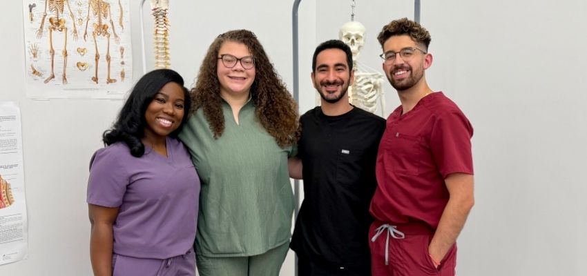 Four students in scrubs pose together in an anatomy lab with a skeleton model behind them.