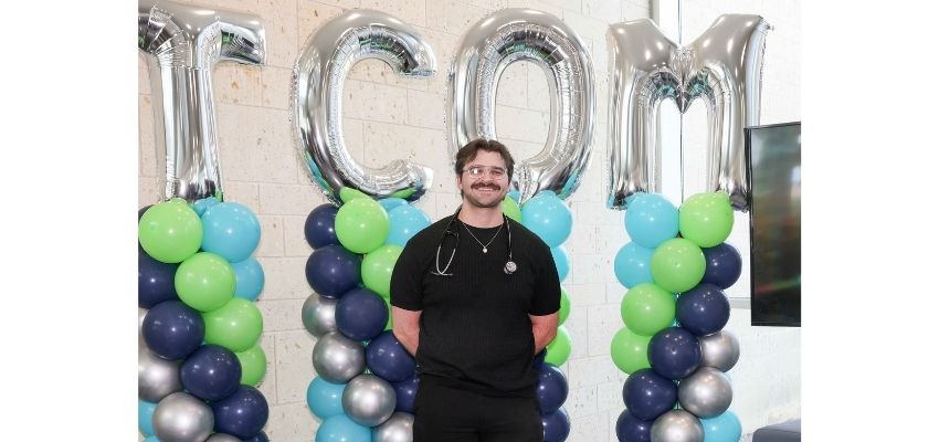 A medical student wearing a stethoscope poses in front of balloon columns and large silver TCOM letters.