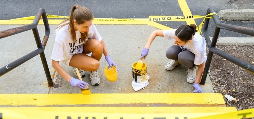 Two people wearing gloves paint the edge of outdoor steps bright yellow as part of a service project.