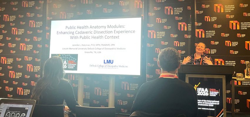 A presenter speaks about public health anatomy modules during an international anatomy conference, with a presentation slide displayed beside the podium.