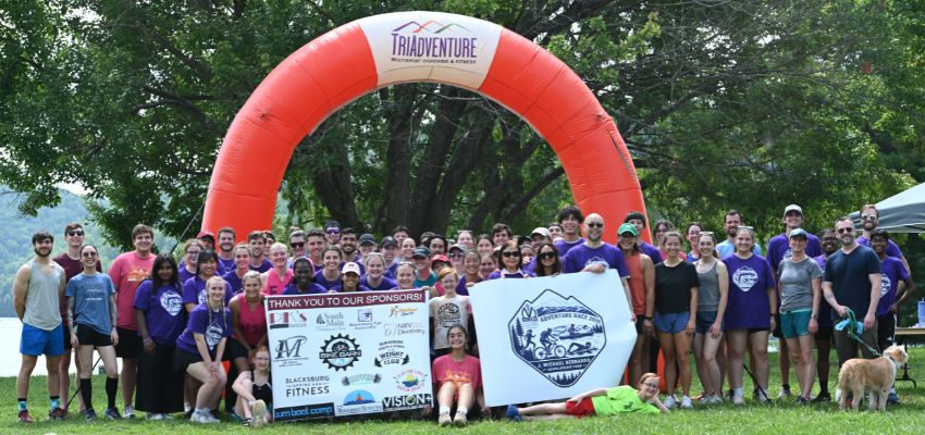 A large group of participants poses outdoors beneath an inflatable arch following an adventure race event.