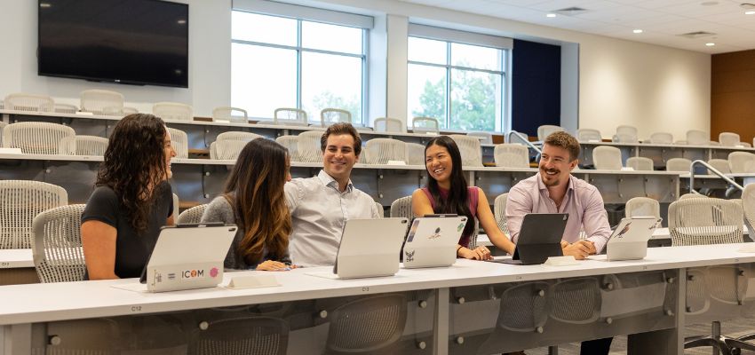 Five students sit together with tablets in a tiered classroom, smiling and talking with one another.