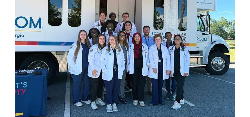 A group of medical students and faculty in white coats poses in front of a mobile medical unit.