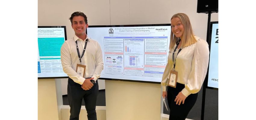 Two students pose beside a digital research poster about AI driven ultrasound image acquisition for echocardiography training.