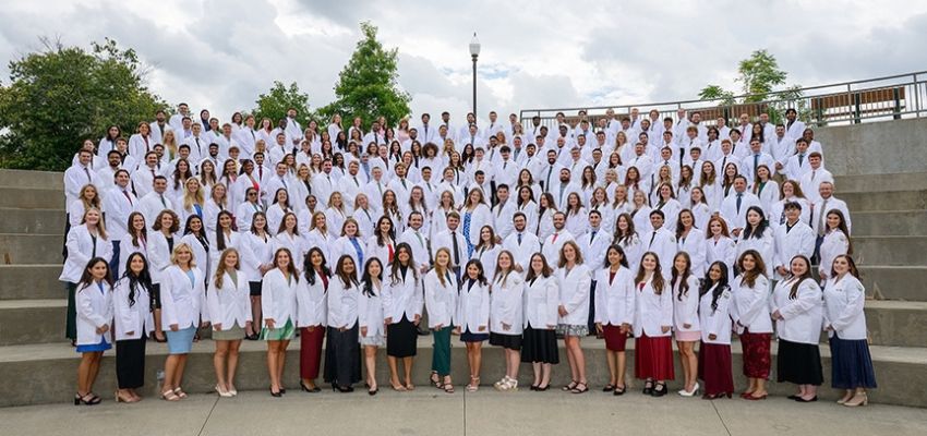 A large group of medical students wearing white coats poses together in an outdoor amphitheater.