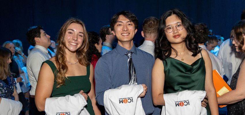 Three students pose together at an event while holding folded white coats.