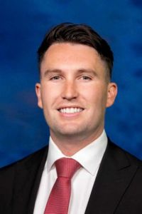 Professional headshot of a smiling man wearing a dark suit and red tie.