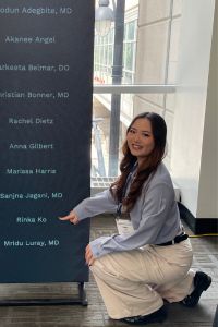 A medical student in a white coat kneels beside a display featuring a list of names and points to her name.