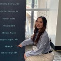 Amedicalstudentinawhitecoatkneelsbesideadisplayfeaturingalistofnamesandpointstohername