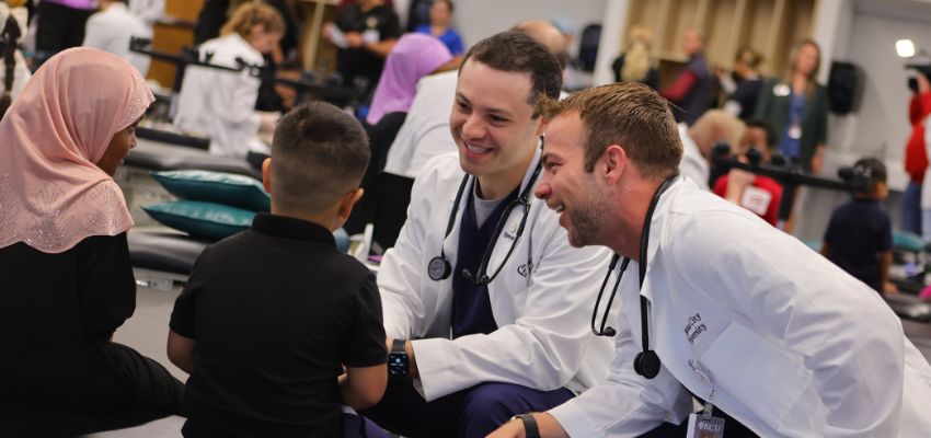 Two medical professionals in white coats speak with a child and an adult during a community health event.
