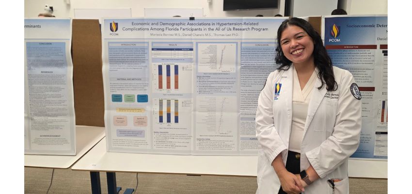 A medical student in a white coat stands beside a research poster on hypertension related complications among Florida participants in the All of Us Research Program.