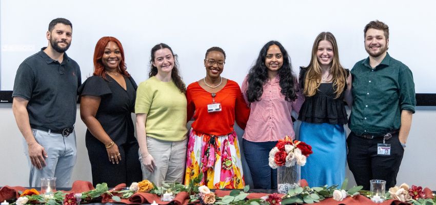 Seven people pose together behind a decorated table with flowers at an indoor event.
