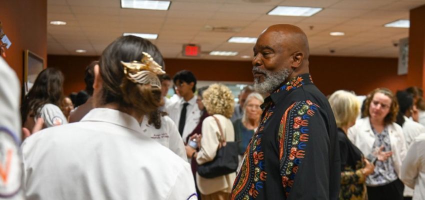 A man in a colorful patterned shirt speaks with medical students and other attendees during a crowded indoor gathering.